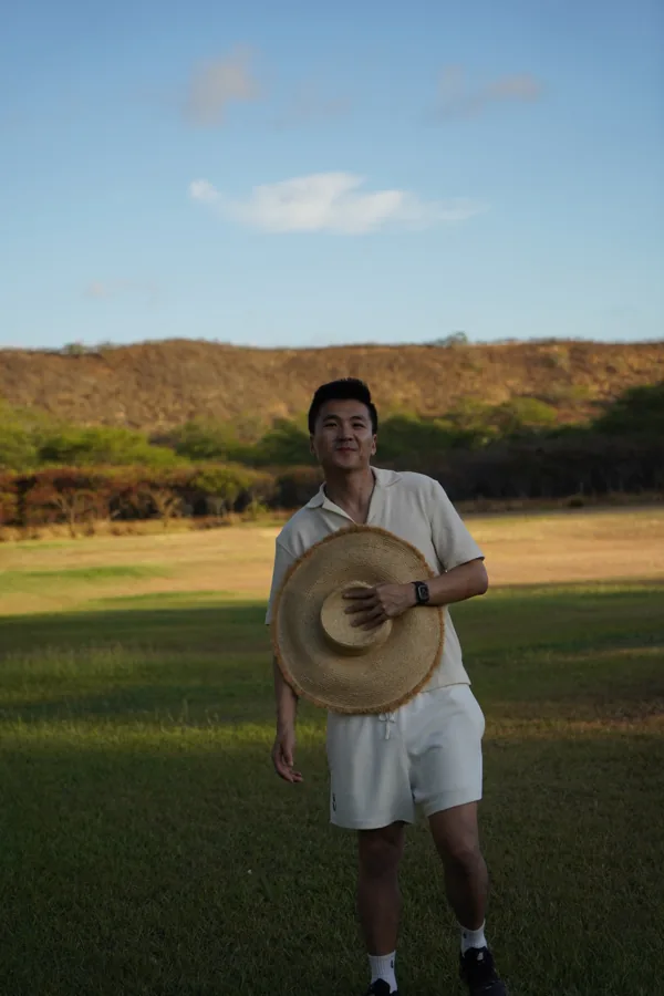 Kelvin standing in a golden field at sunset in Hawaii, holding a hat